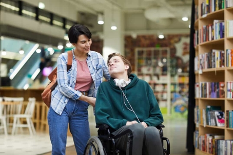 Boy in wheelchair with headphones around his neck looks up at a woman holding onto his wheelchair in a library